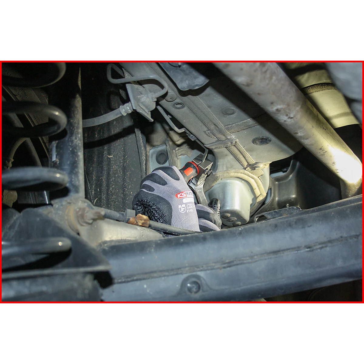 Nahaufnahme von jemandem, der unter einem Auto arbeitet. Eine Hand in einem grauen Handschuh hält ein Werkzeug.